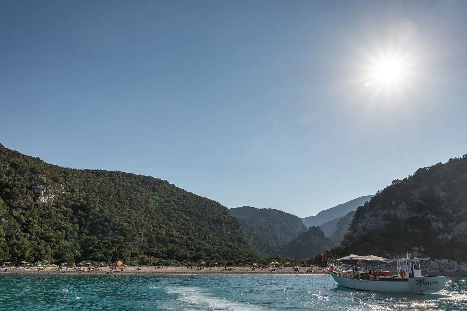 Una barca da escursione davanti alla spiaggia di Cala Luna, con i monti coperti di macchia e il sole alto nel cielo