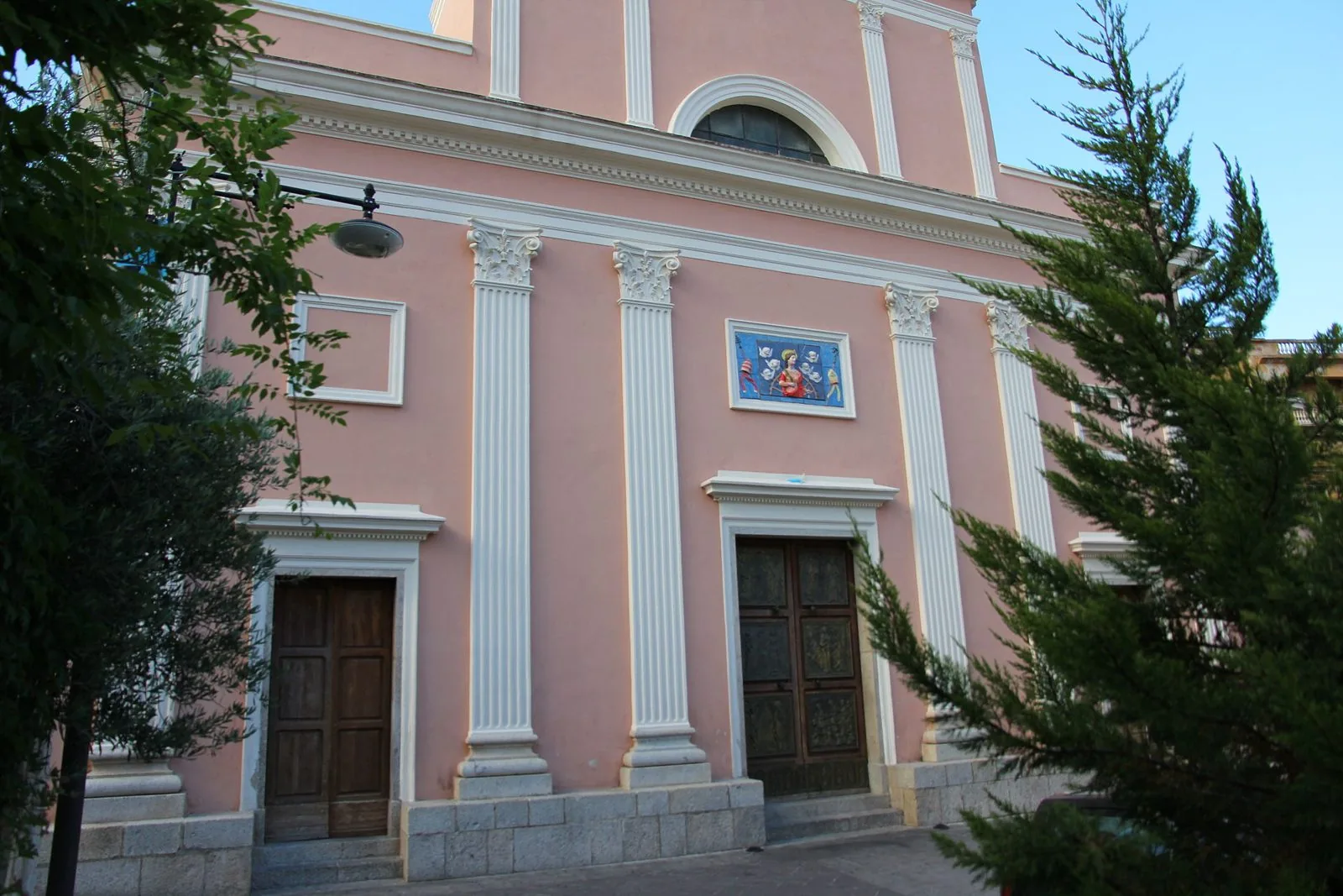 Facciata rosa con lesene bianche della chiesa di Santa Caterina d'Alessandria a Dorgali, con il pannello in ceramica sopra il portale