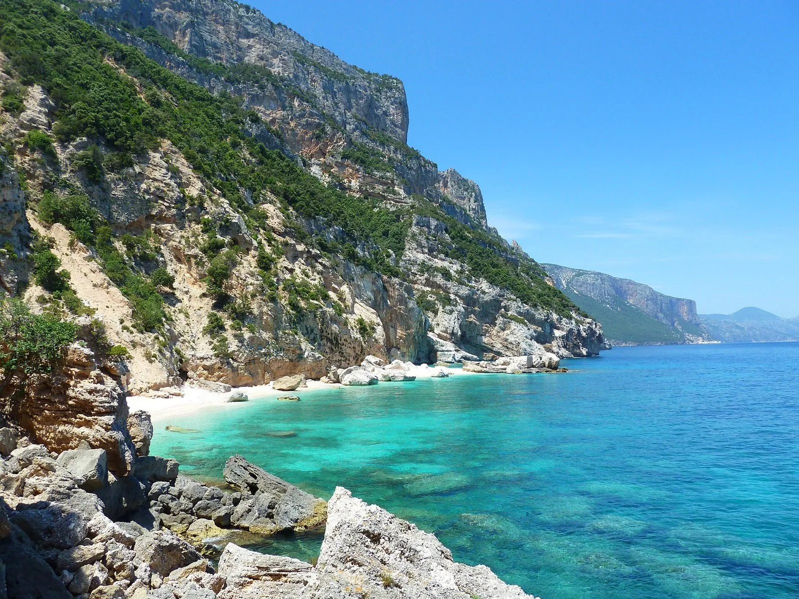 Cala con spiaggia bianca e acqua turchese ai piedi di alte pareti calcaree coperte di macchia mediterranea